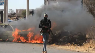 أحد أفراد قوات الأمن السورية يمرّ أمام سيارة محترقة في منطقة المزرعة، قرب السويداء