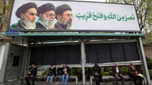  إيرانيون يجلسون في محطة حافلات أسفل لوحة إعلانية للنظام