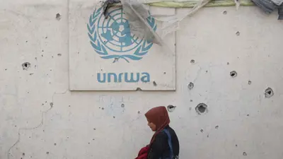 امرأة تسير بجانب سور عليه شعار الأونروا