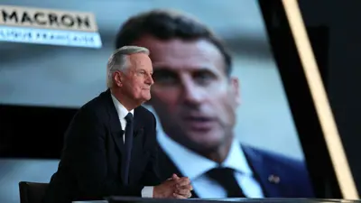 بارنييه خلال لقاء تلفزيوني وصورة ماكرون في الخلفية